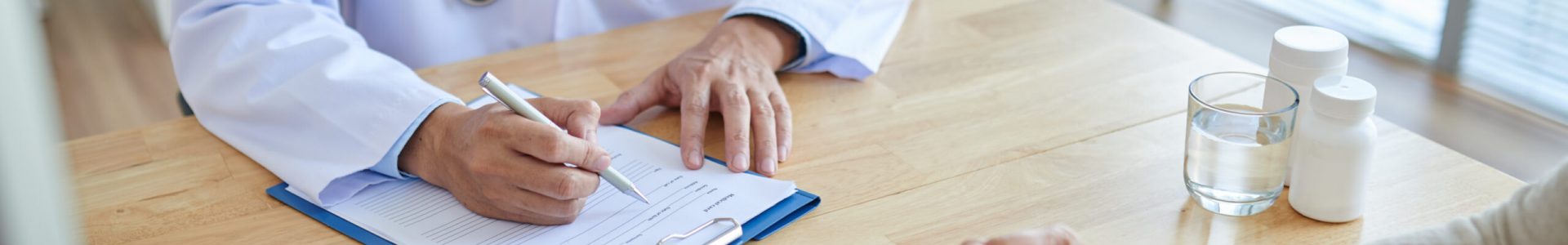 Close-up shot of male doctor filling in medical record, his patient sitting opposite him